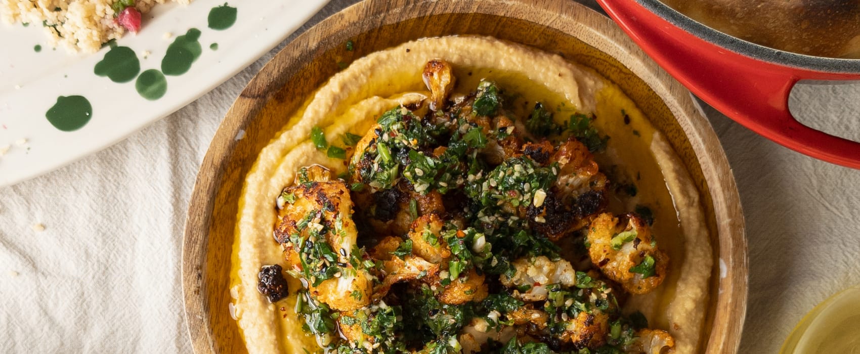 Eine Schüssel mit Hummus, darauf gerösteter Blumenkohl und Gremolata. Daneben Couscoussalat und Fladenbrot.
