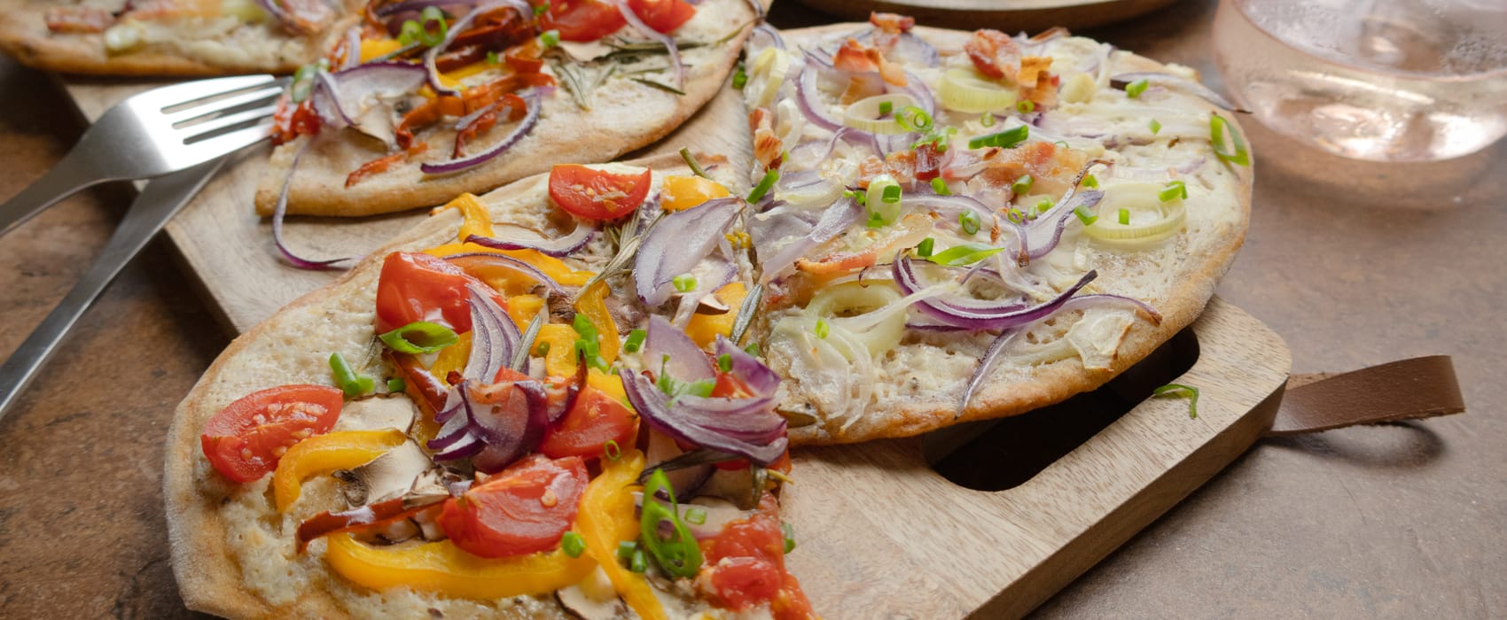 Zweierlei Flammkuchen belegt mit Gemüse und Speck auf Holzbrett angerichtet. 