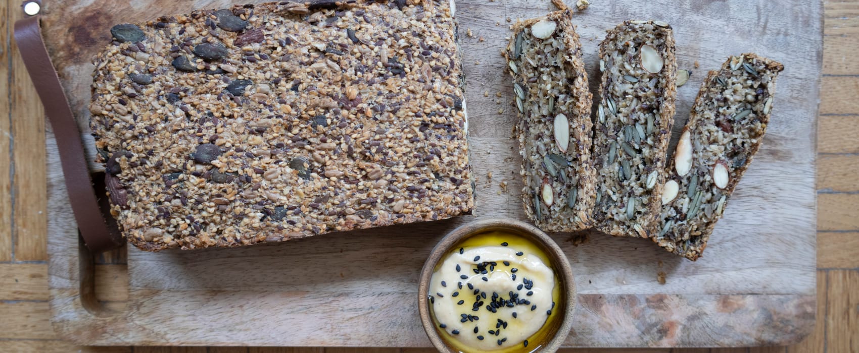 Teilweise aufgeschnittenes Saatenbrot auf Holzbrett, dazu ein Schälchen mit Hummus